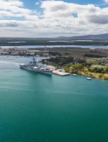 An aerial view of Pearl Harbor, featuring a historic battleship docked near lush greenery and calm blue waters.
