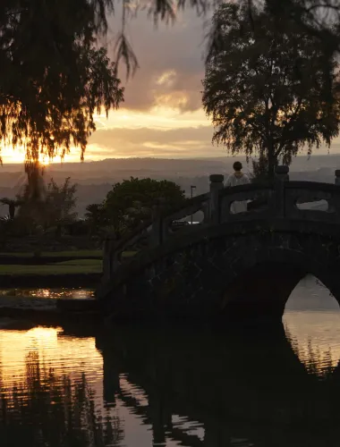 A scenic view of Liliuokalani Gardens, a Japanese garden, at sunset with a traditional arched bridge over a calm pond.