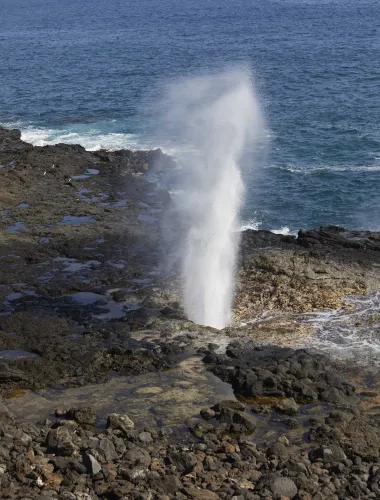 Spouting Horn blowhole erupting water into the air against a rocky shoreline.