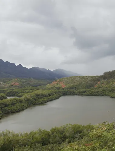 A serene fishpond surrounded by lush green mountains under a cloudy sky.