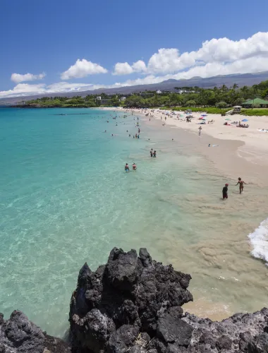 Scenic image of Hapuna Beach State Park with turquoise water, white sand, and distant mountains, surrounded by lush greenery.