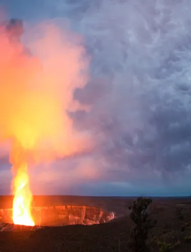 Glowing lava erupts from a volcanic crater, lighting up the dark sky with vibrant orange hues against dramatic storm clouds.