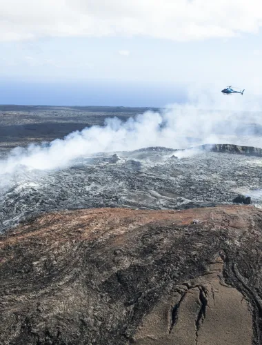 Aerial view of a volcanic landscape with active lava flows and a helicopter in the distance.