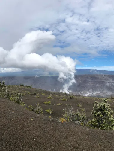 A landscape of Hawai‘i Volcanoes National Park featuring a steaming volcanic crater surrounded by barren terrain and sparse greenery under a cloudy sky.