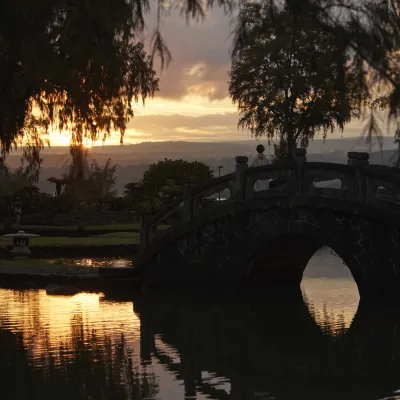 A scenic view of Liliuokalani Gardens, a Japanese garden, at sunset with a traditional arched bridge over a calm pond.