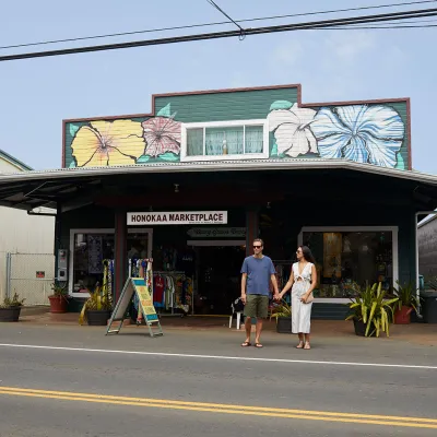 Couple holding hands outside the Honokaa Marketplace, a colorful building decorated with hibiscus murals, on a sunny day.