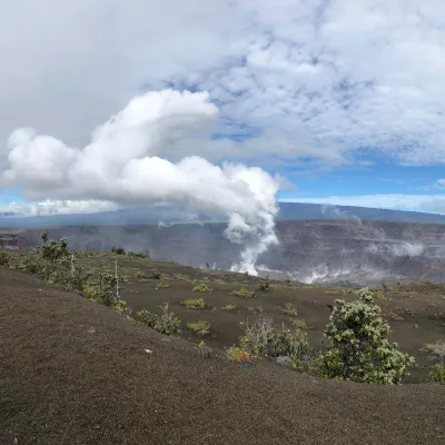 A landscape of Hawai‘i Volcanoes National Park featuring a steaming volcanic crater surrounded by barren terrain and sparse greenery under a cloudy sky.