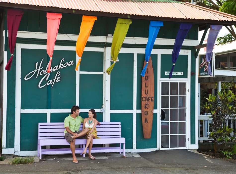 Couple sitting on a purple bench outside the Holualoa Cafe, a charming coffee shop adorned with colorful flags and a surfboard sign.
