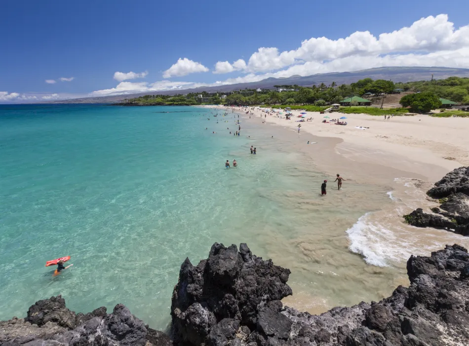 Visitors enjoying the turquoise waters and white sands of Hapuna Beach State Park with a rocky volcanic coastline in the foreground.