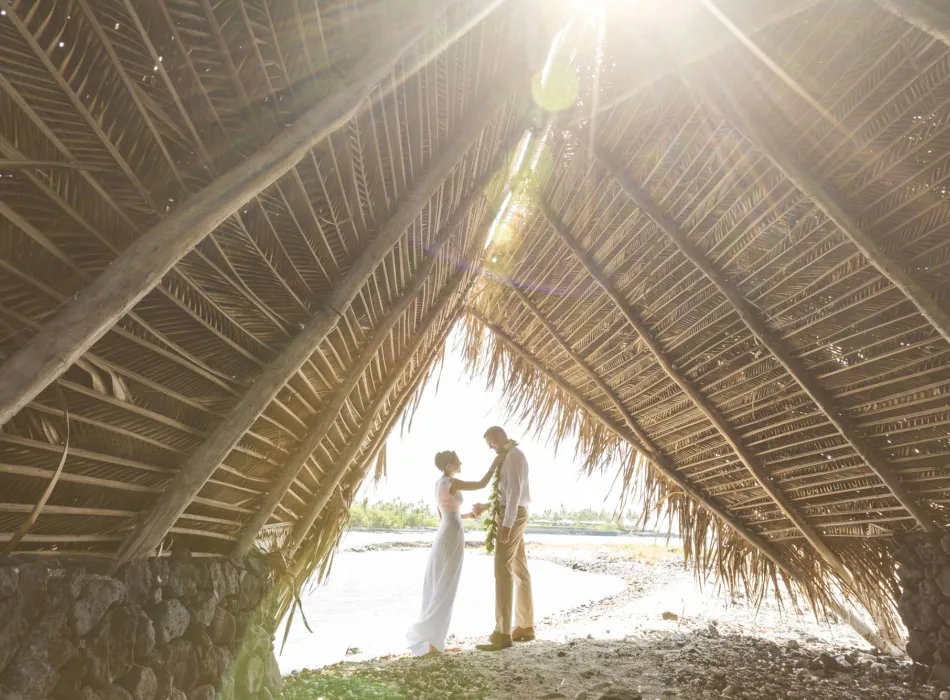 A couple stands under a traditional palm-frond structure by the water, bathed in sunlight.