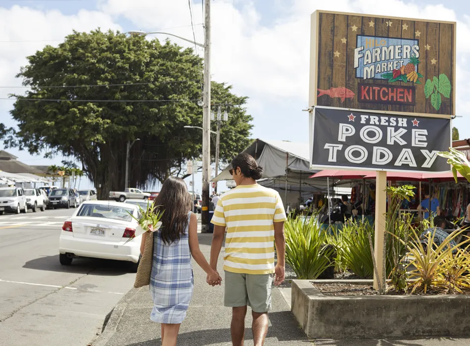 Couple walking toward a market with a sign reading 'Fresh Poke Today' and stalls selling local goods and food in the background.