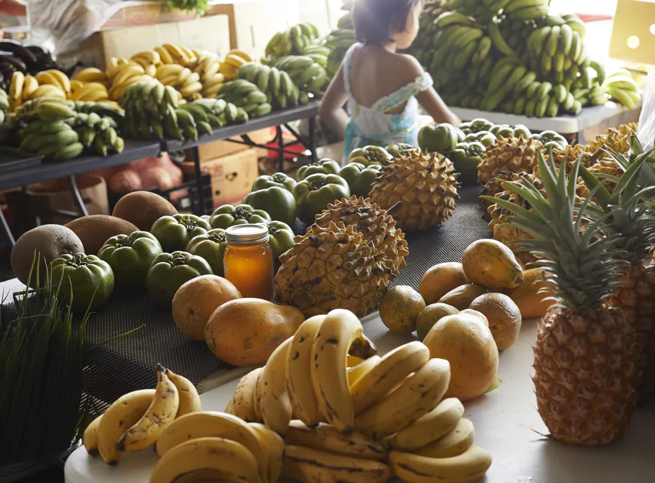 Table filled with tropical fruits such as bananas, pineapples, and papayas at the Hilo Farmers Market, with a child in the background.