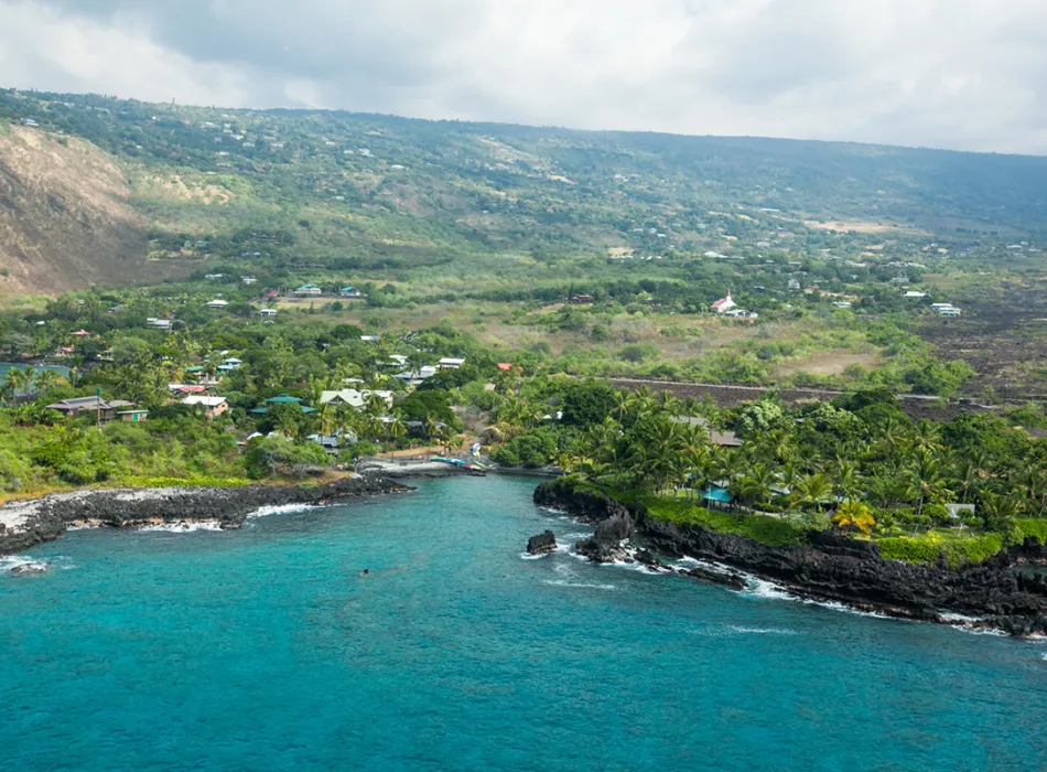 Aerial view of Kealakekua Bay with turquoise waters, lush greenery, rocky cliffs, and scattered buildings along the shoreline.