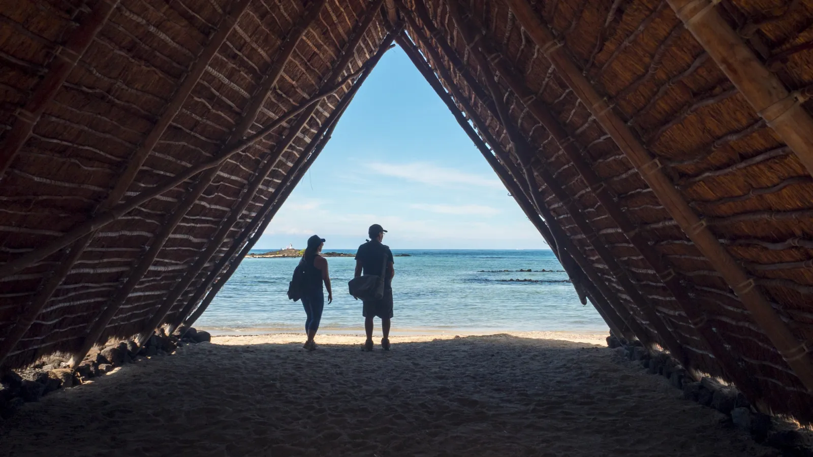 Two people walking under a traditional Hawaiian thatched hale with the ocean in the background.